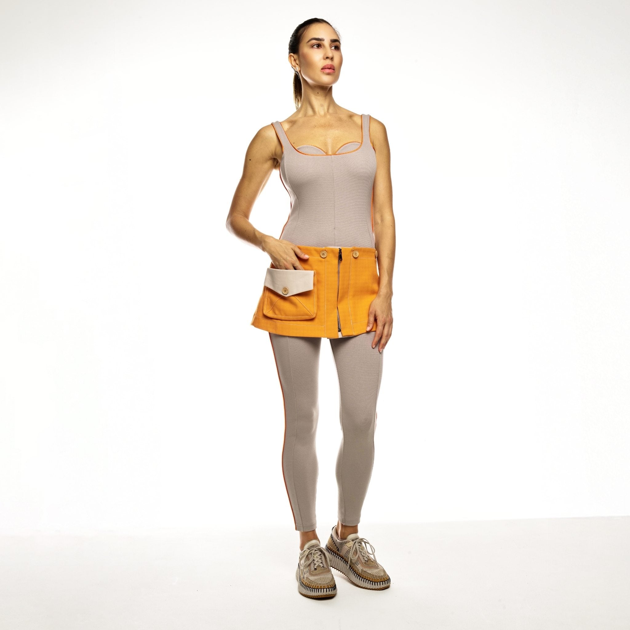 Woman wearing a beige jumpsuit, orange skirt, and grey leggings on a white background